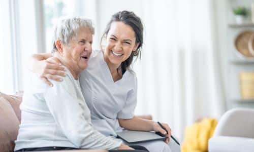 Lachende Pflegerin umarmt eine ältere Person, die im Rollstuhl sitzt.