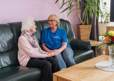 Zwei Frauen, eine Pflegekraft und eine ältere Bewohnerin, halten Hände und unterhalten sich in der Wohngemeinschaft
