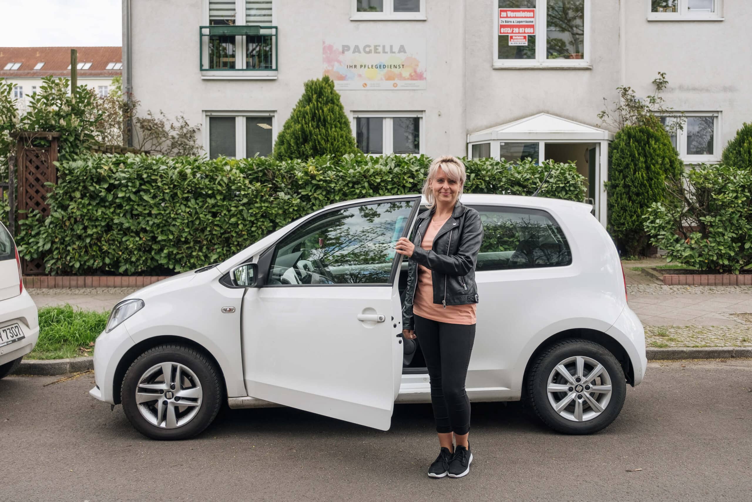 Eine Pflegefachkraft von Pagella steht neben einem weißen Auto vor dem Gebäude des Pflegedienstes.
