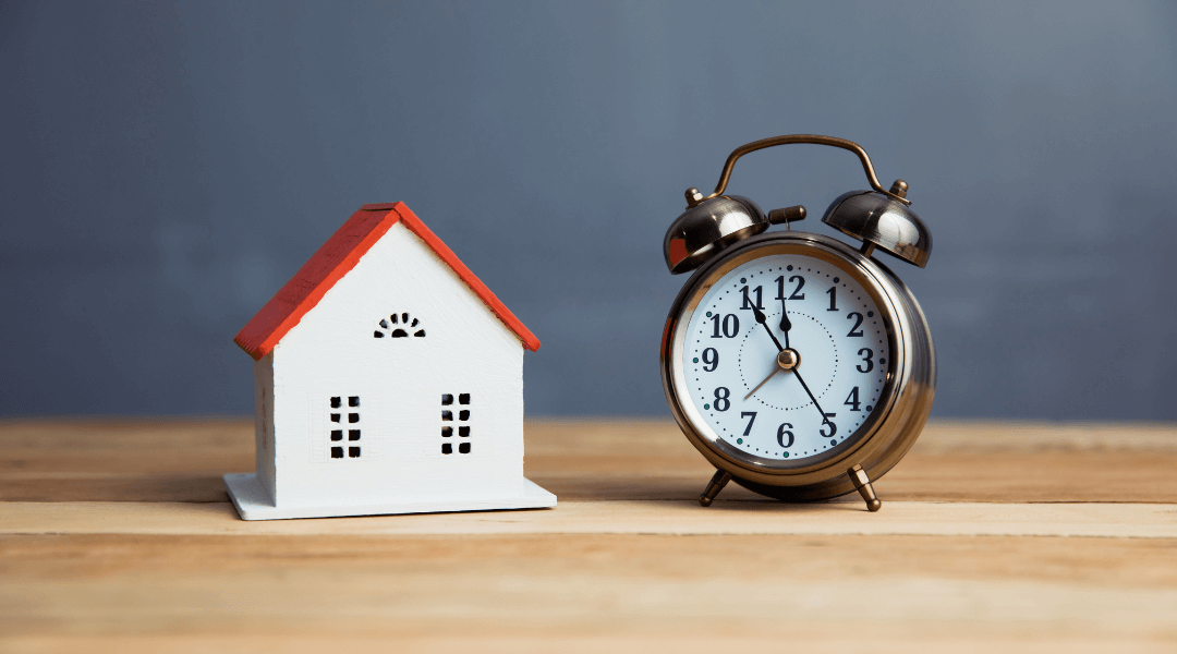 Ein kleines Modellhaus und ein Wecker stehen nebeneinander auf einem Holztisch.