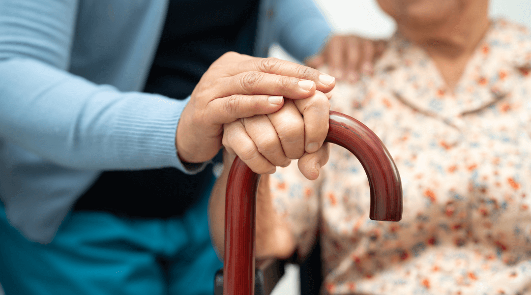 Nahaufnahme von unterstützenden Händen, die einer älteren Person mit Gehstock helfen.