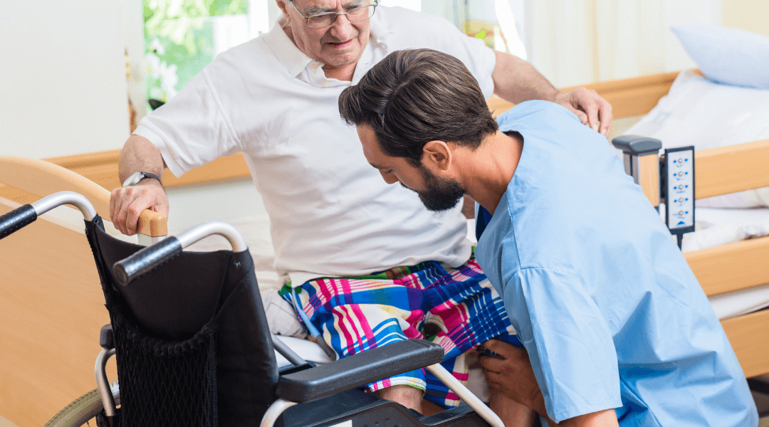 Ein Pflegehelfer hilft einem Senior beim Aufstehen aus dem Rollstuhl.